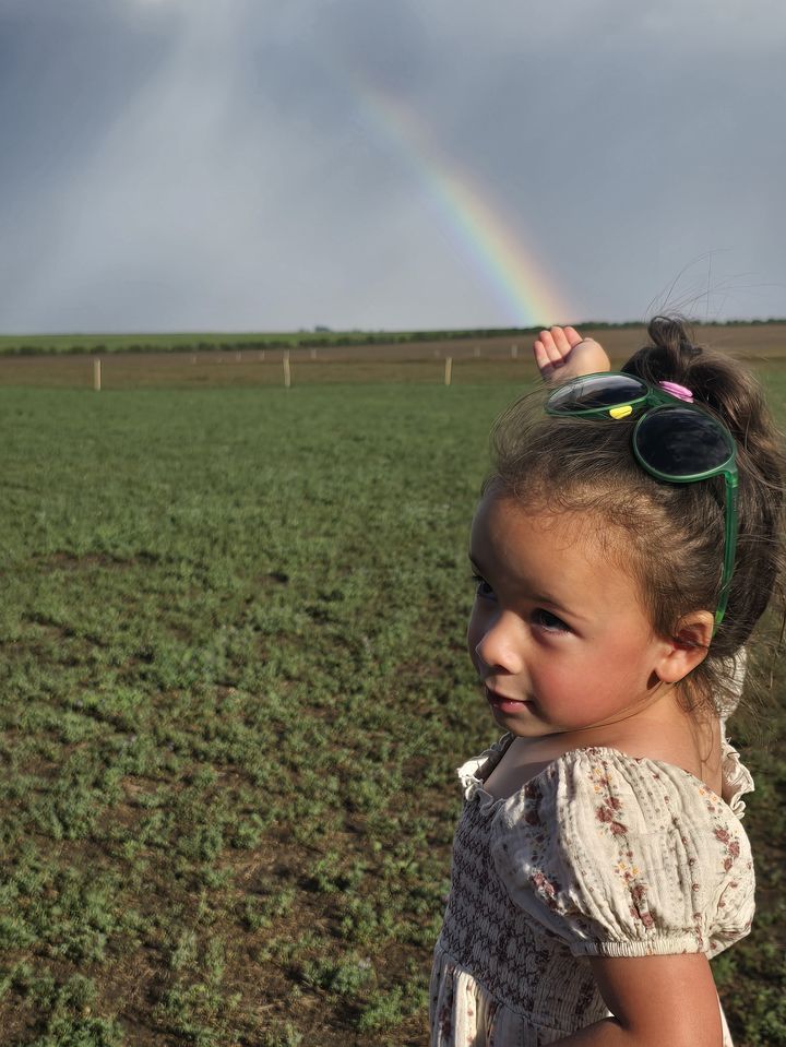 A young girl in a floral dress on the farm, pointing up at a rainbow over the green field