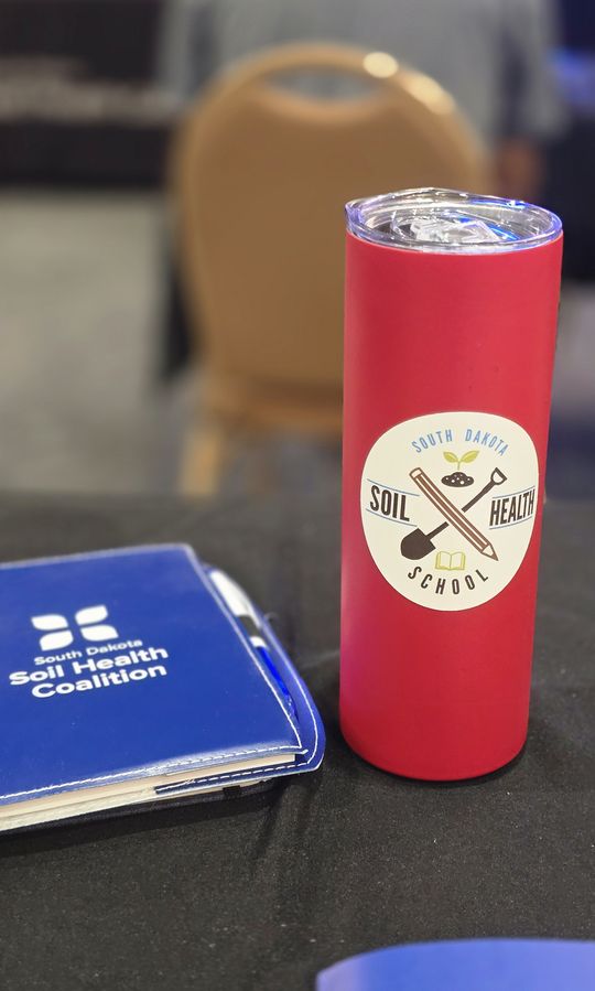 A red South Dakota Soil Health School tumbler beside a South Dakota Soil Health Coalition notebook