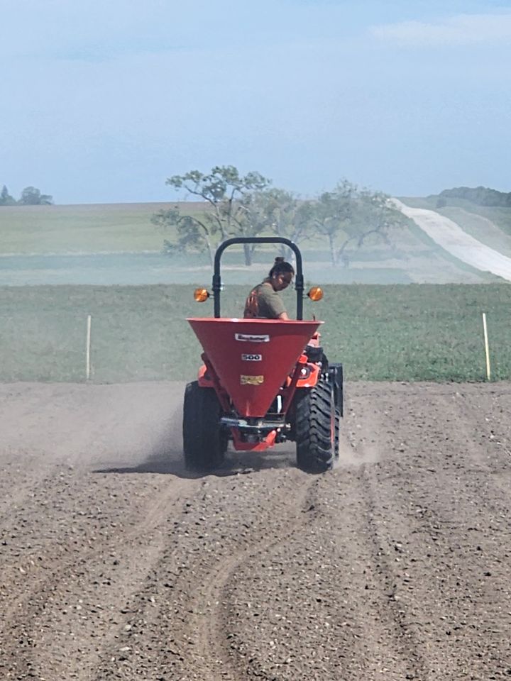 Sarah on an orange tractor, planting cover crop with a red seed spreader across a prepared field