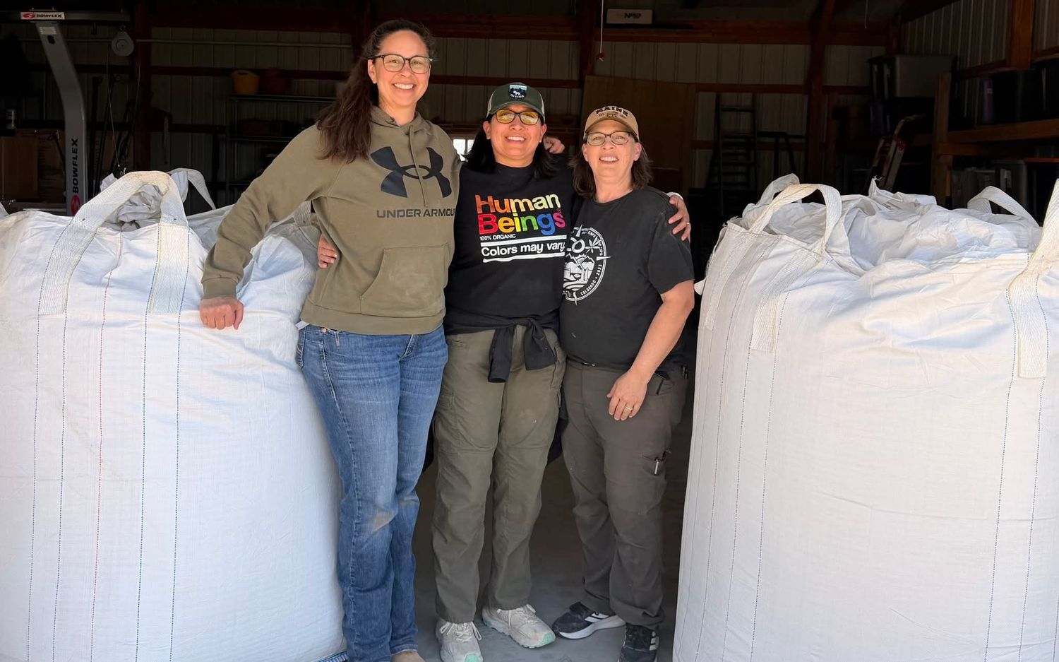 Sarah Downey, Dionne Holmquist and Nickie Stowell, left to right, smiling together in the barn between two large bulk bags